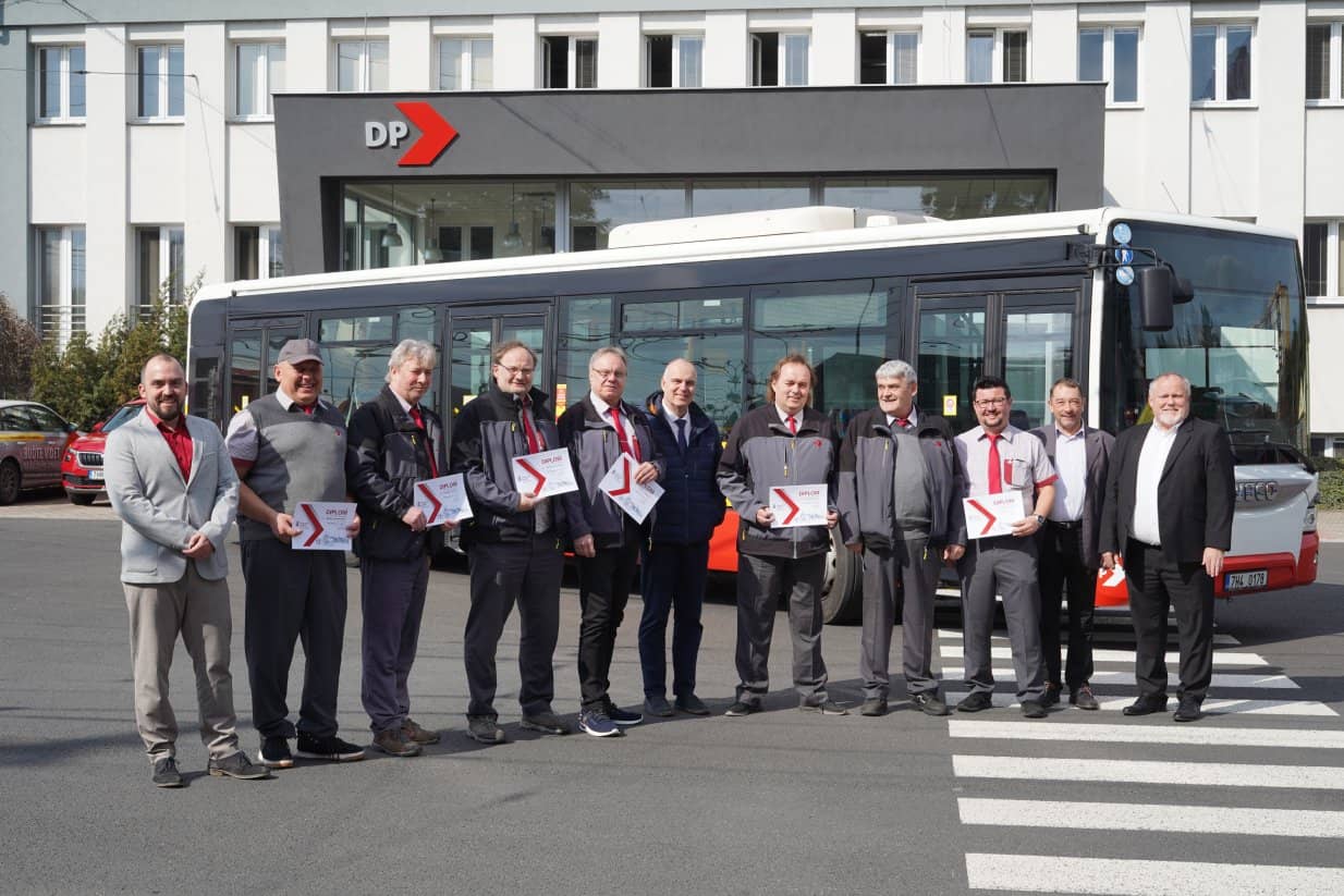 Group photo of eleven men standing at a crosswalk in front of a large white bus and a building with a „DP“ logo. Most of the men are wearing public transport company uniforms (gray jackets or vests with red details) and are holding white diplomas with a red arrow symbol in their hands. On the sides, men in civilian clothes and suits are standing.
