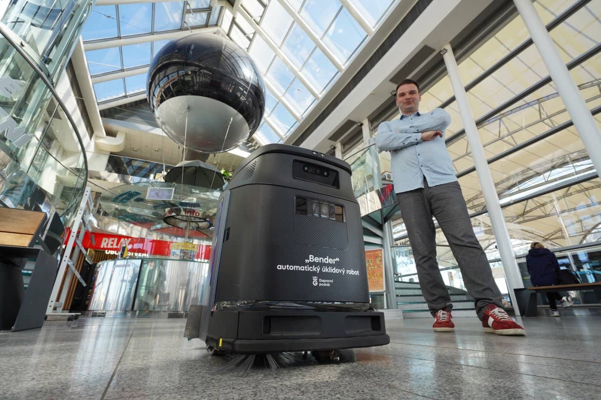 Pohled z nízkého úhlu na černého automatického úklidového robota s nápisem „Bender automaticý úklidový robot – Dopravní podnik“. Vedle robota stojí muž s překříženýma rukama v košili a teniskách. Nacházejí se v moderní prosklené hale s vysokým stropem, kde v pozadí dominuje velká černá dekorativní koule zavěšená v prostoru a prodejna Relay.