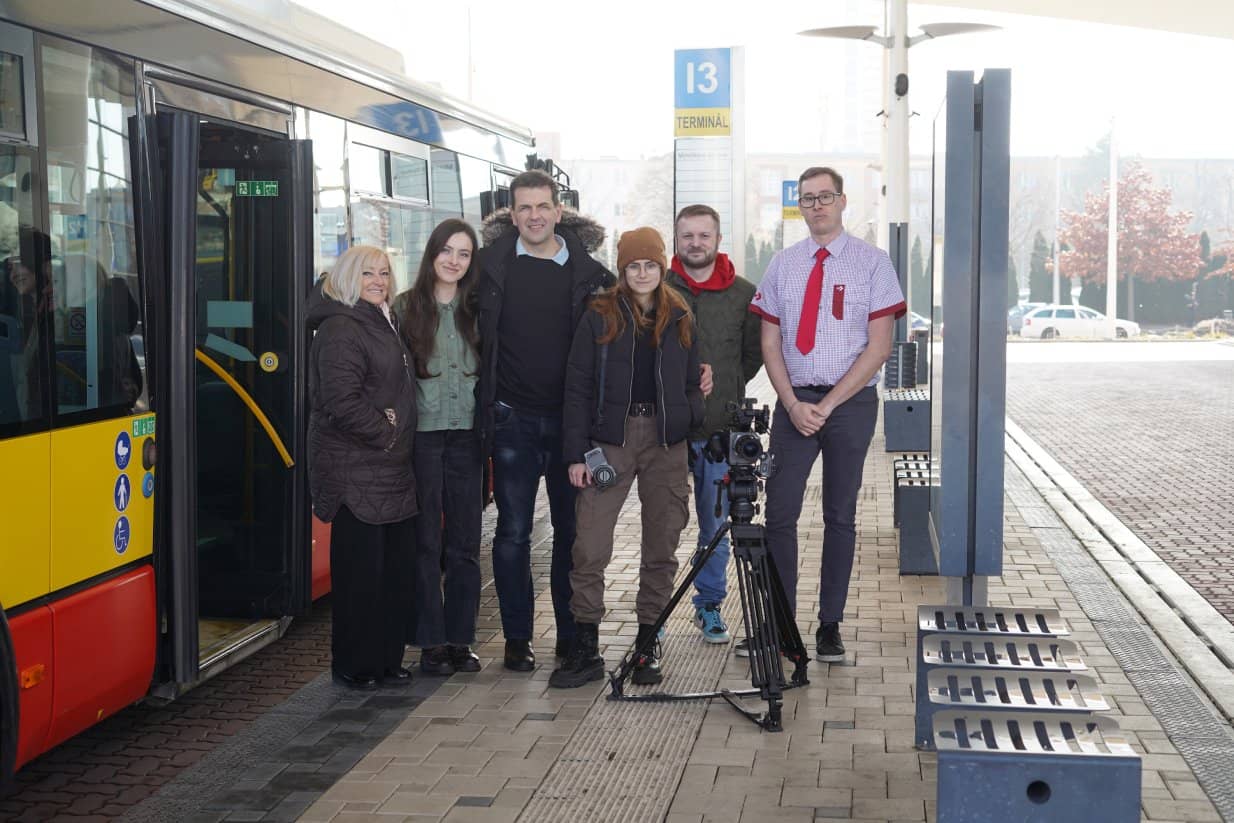 Skupina šesti lidí pózuje na autobusovém terminálu vedle moderního červeno-žlutého autobusu. Mezi nimi stojí profesionální kamera na stativu. V pozadí je vidět označník s číslem terminálu 13 a budovy v mlhavém oparu. Lidé jsou oblečeni v civilním oblečení (zimní bundy, čepice), jeden muž na pravém kraji má na sobě uniformu řidiče – košili s červenou kravatou.