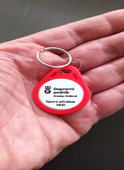 Outstretched palm with a chip pendant lying on it. It forms a white circle with the inscription Dopravní podnik Hradec Králové, a public transport chip pendant, which is set in a red casing.