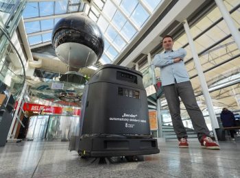 Pohled z nízkého úhlu na černého automatického úklidového robota s nápisem „Bender automaticý úklidový robot – Dopravní podnik“. Vedle robota stojí muž s překříženýma rukama v košili a teniskách. Nacházejí se v moderní prosklené hale s vysokým stropem, kde v pozadí dominuje velká černá dekorativní koule zavěšená v prostoru a prodejna Relay.